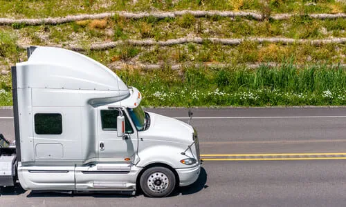 A white semi trailer truck driving along a road with a terraced hill in the background.