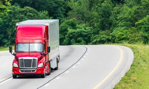 A red big rig truck and its trailer delivering cargo along a road with thick vegetation in the background.