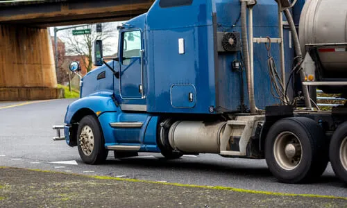 A blue big rig truck transporting a fuel tank trailer and about to make a turn under a bridge.