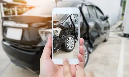 An accident victim documenting damage to her black car on her smartphone in the afternoon.