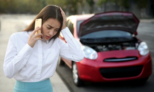 A woman calling for help on her phone after her red car was damaged in an accident.