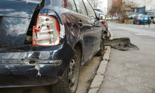 A dark blue car damaged in an accident stopped along the side of a city road.