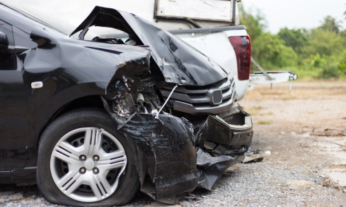 A black car in a parking lot awaiting repairs after a collision with a pickup truck.